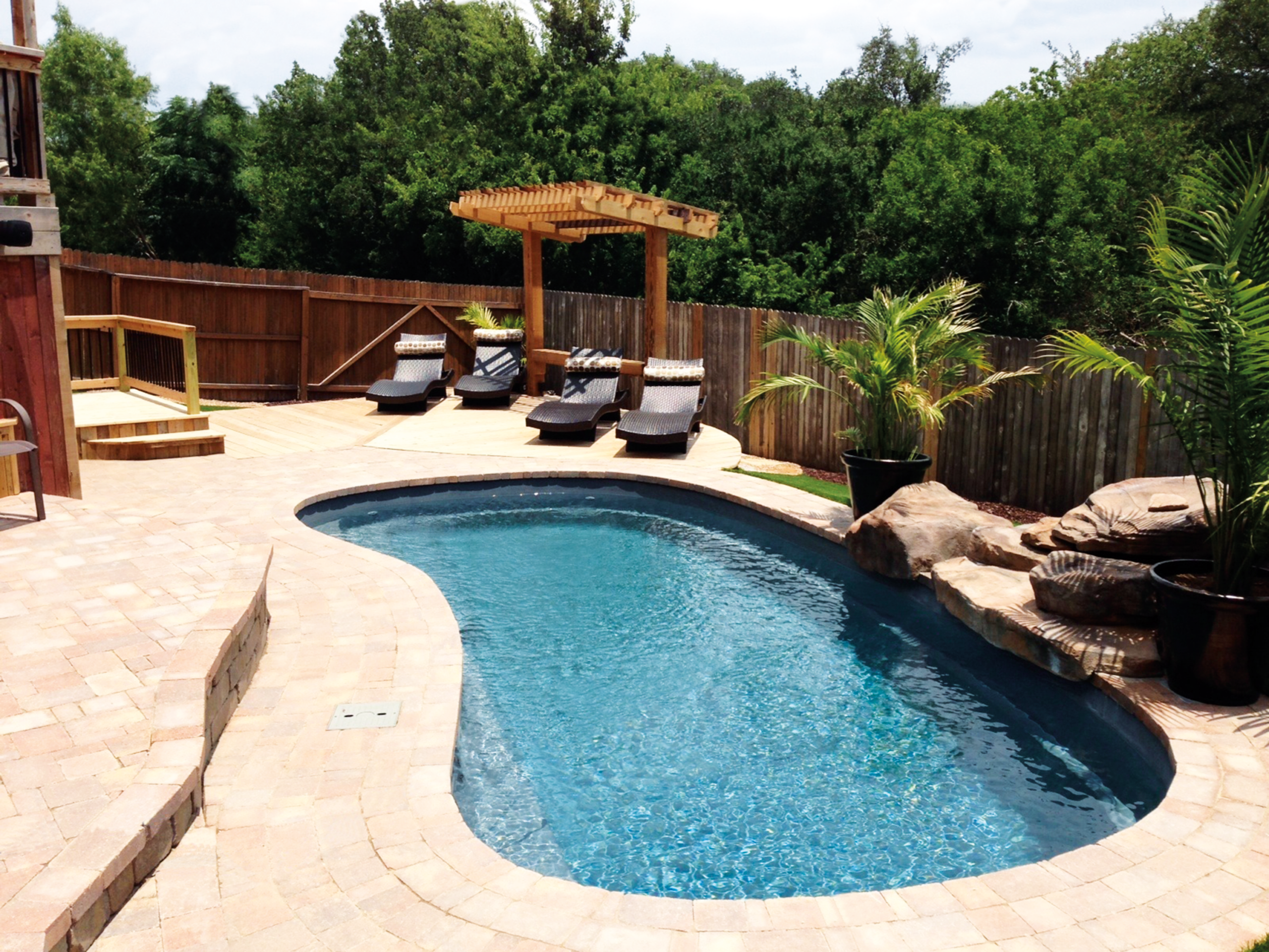 Curved pool with a dark blue interior, surrounded by light-colored paving stones, featuring a rock waterfall, a wooden pergola over lounge chairs, and a privacy fence in the background.