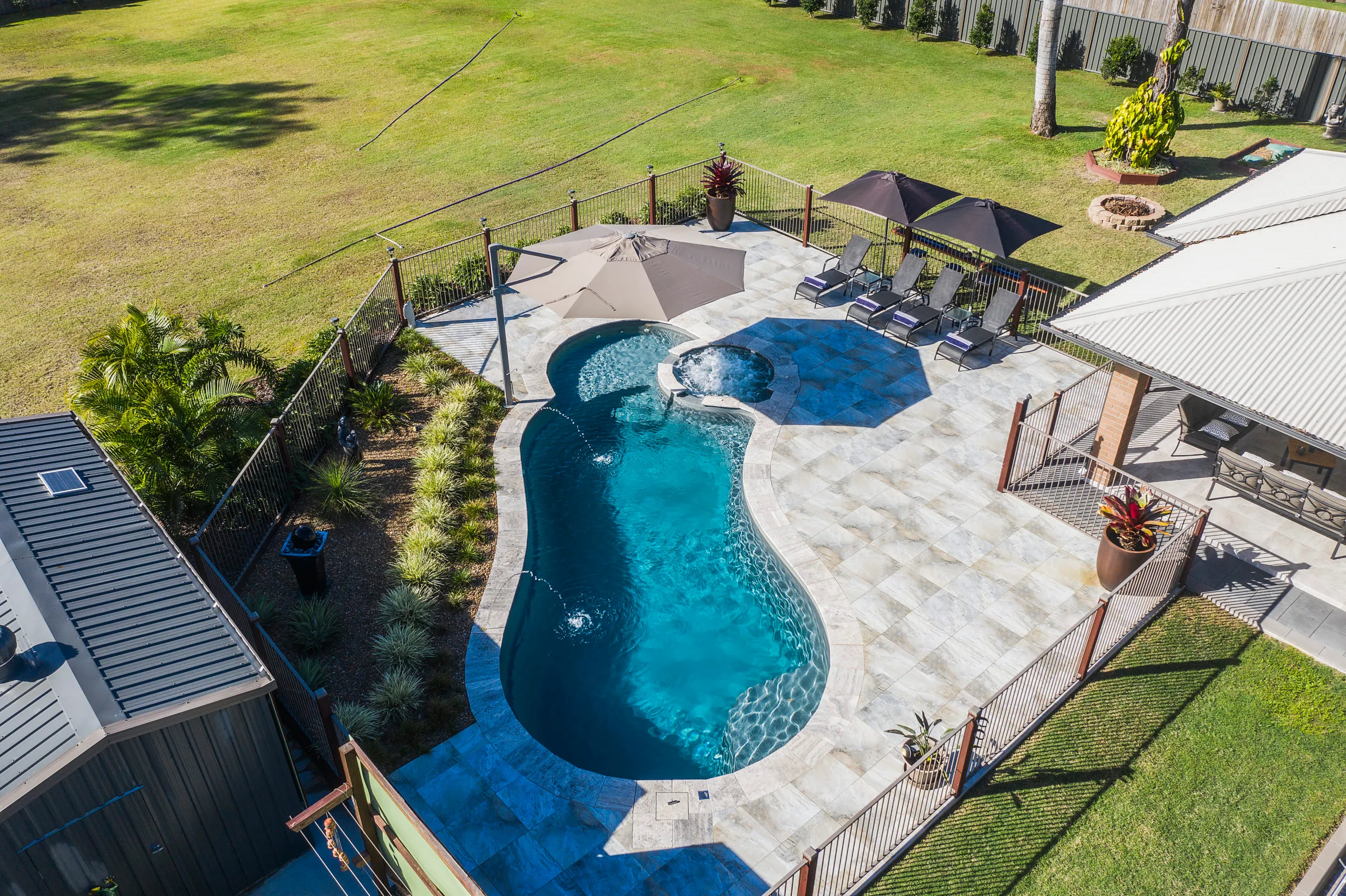 Aerial view of a kidney-shaped fibreglass pool and integrated spa, surrounded by light-colored tile paving and lounge areas under umbrellas, set in a large grassy backyard.