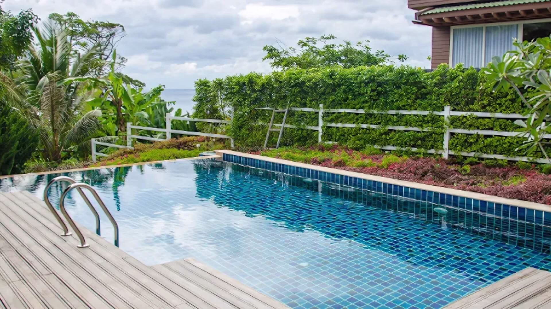Tropical infinity pool with small blue mosaic tiles, a wooden deck, stainless steel ladder, and a white fence overlooking the ocean.