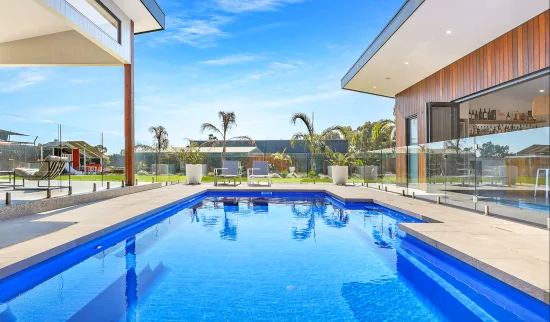 Modern rectangular fibreglass pool with bright blue interior, wide perimeter coping, and surrounding glass fence separating it from a contemporary home and lawn.