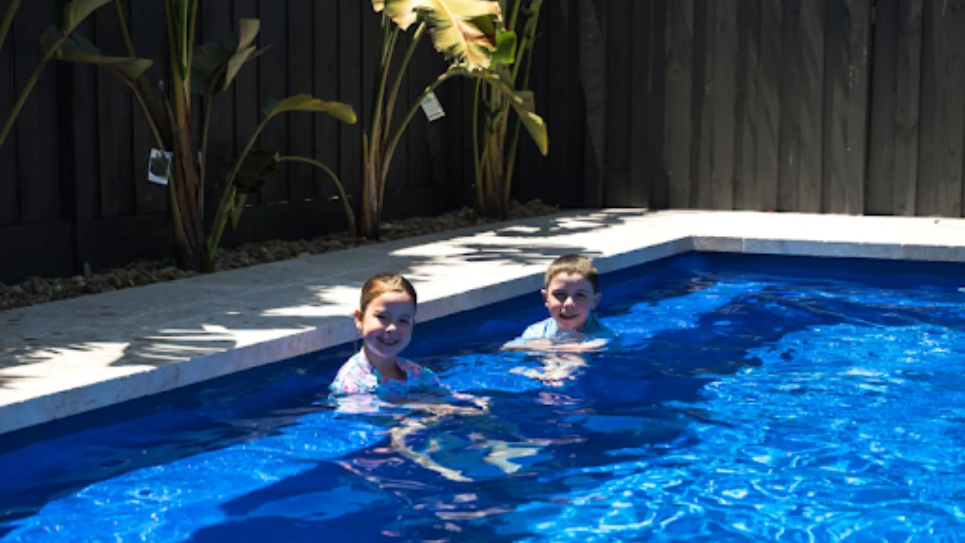 Two children swimming in a fibreglass pool with clear blue water, surrounded by plants and a dark wooden fence.
